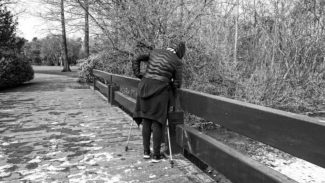 Person walking in a coat with sticks and looking over the side of a bridge