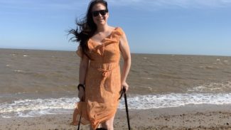 Zoe wears an orange dress and a dog sits in front of her. She's stood on the beach using a black walking stick with the sea behind her