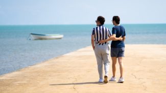 Two men walk on a dock with their arms around each other while facing the sea