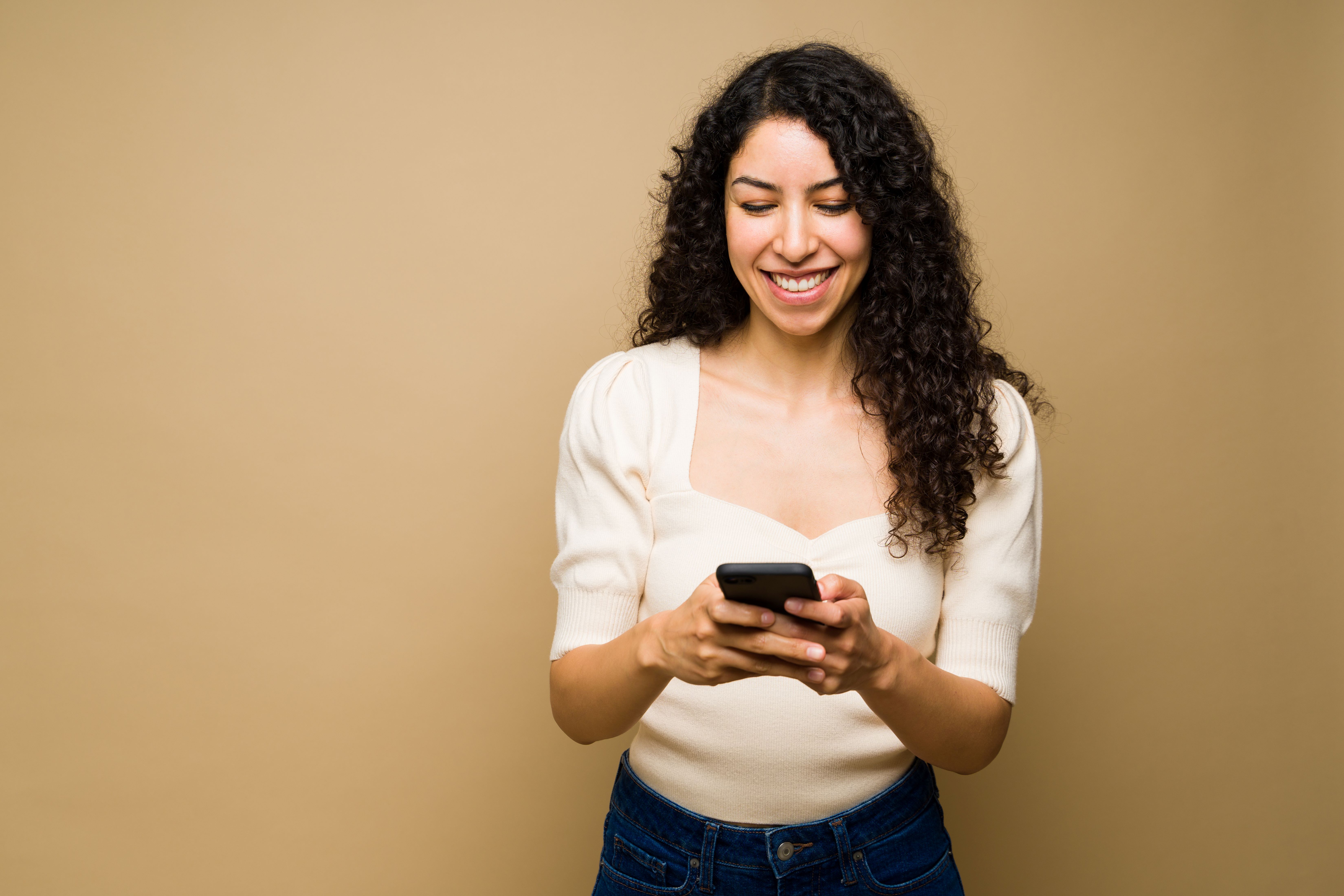 A woman with brown curly hair looks down at her phone while smiling