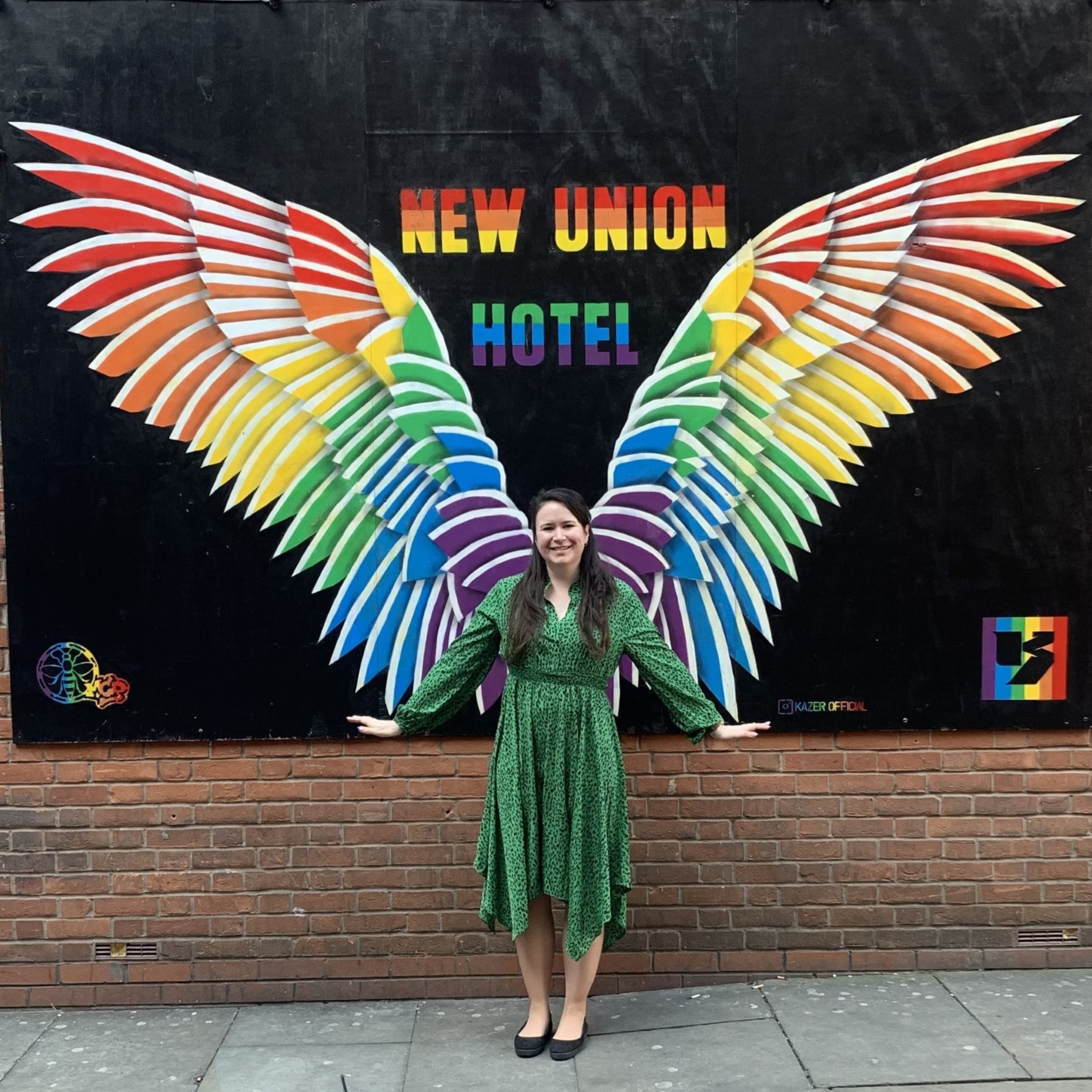 Zoe stands smiling in a green and black dress. Her arms are out to the side and the wall behind her has a mural of rainbow wings
