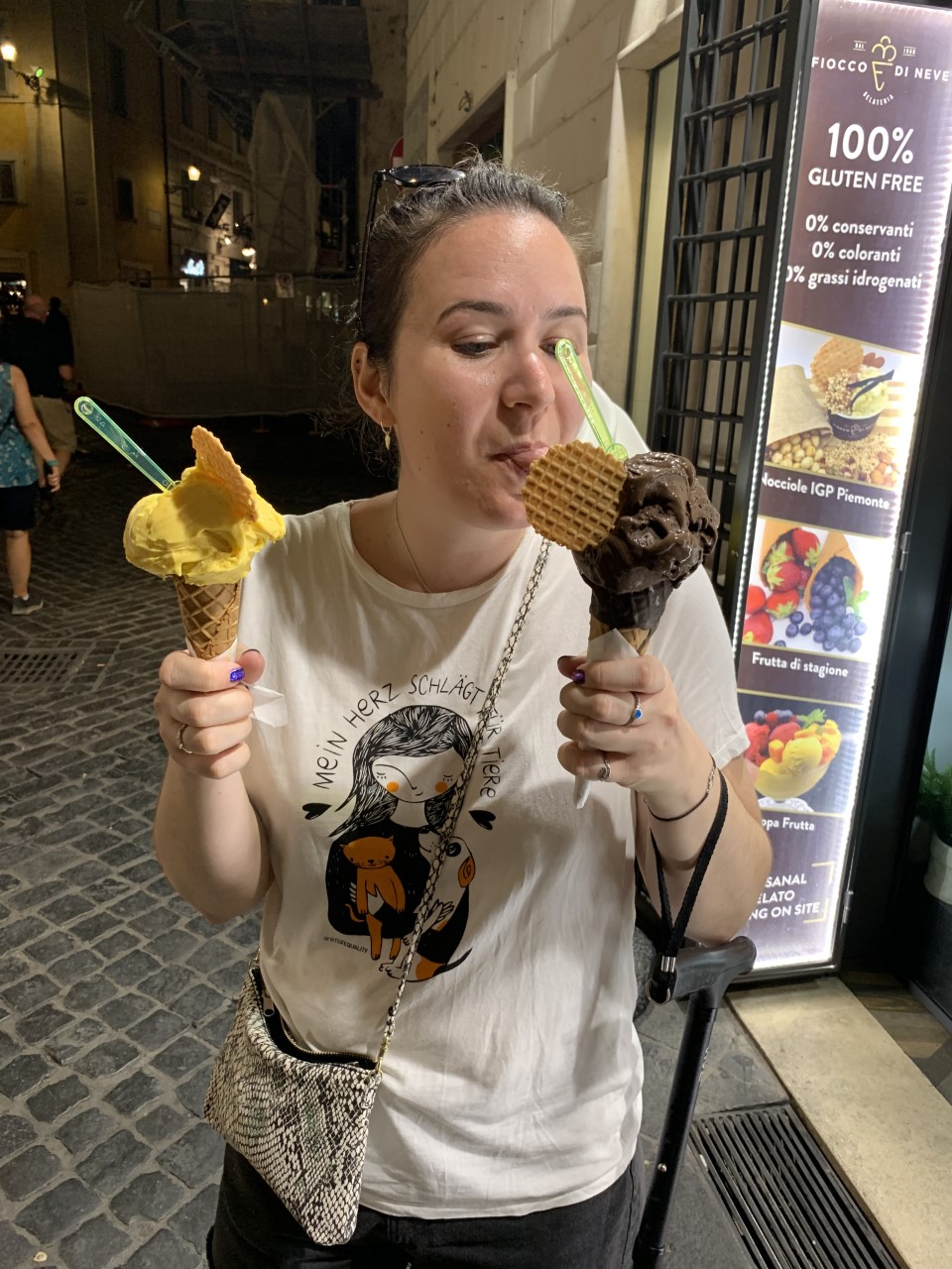 Zoe stands and holds two ice creams, eating one while her stick is attached to her wrist