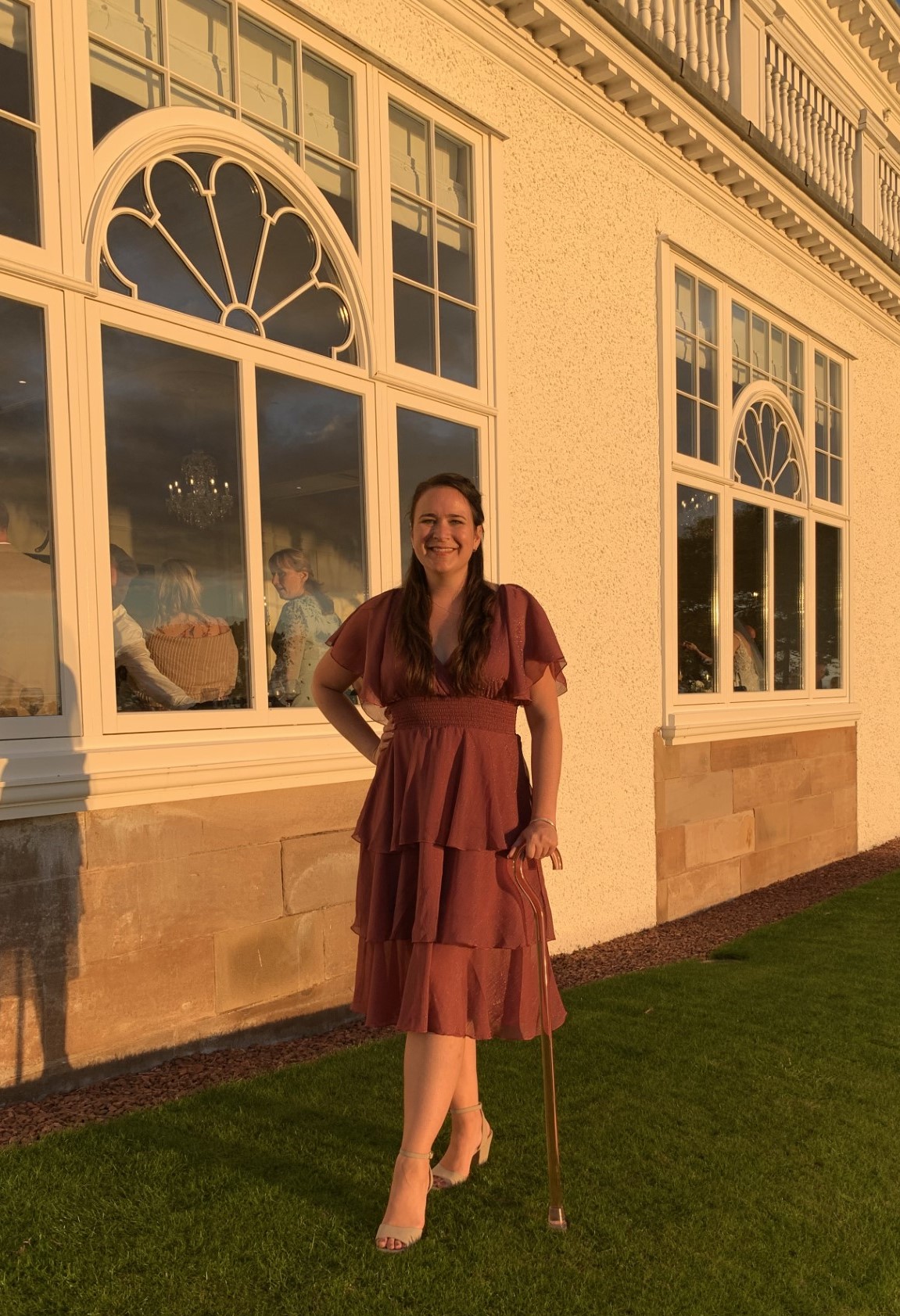 Zoe stands on grass in front of a white building wearing a pink dress and holding a clear pink stick