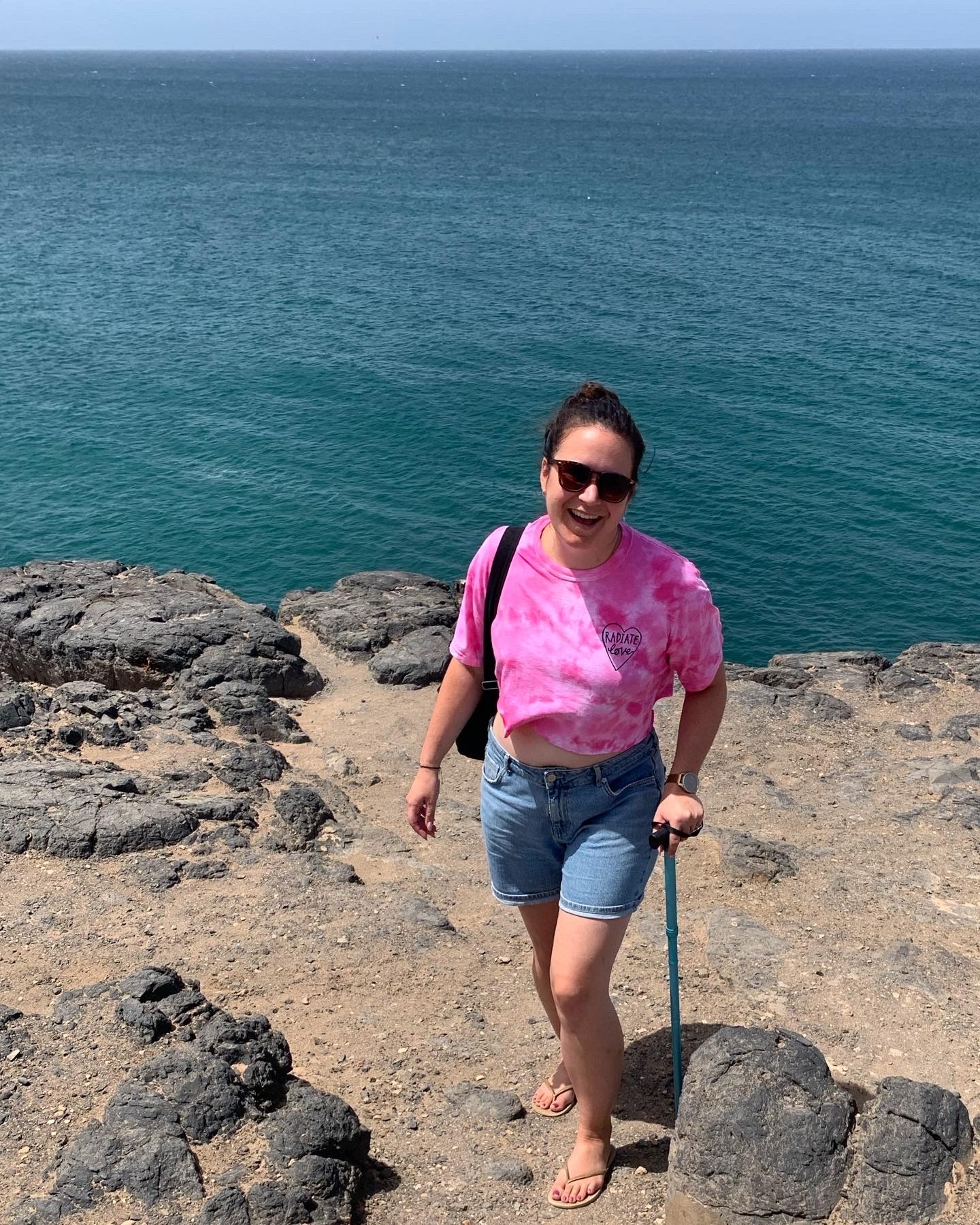 Zoe stands on rocks near the sea, smiling and holding a black walking stick